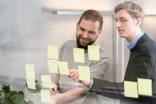 Zwei Personen, die gelbe Haftnotizen an eine Glaswand kleben.